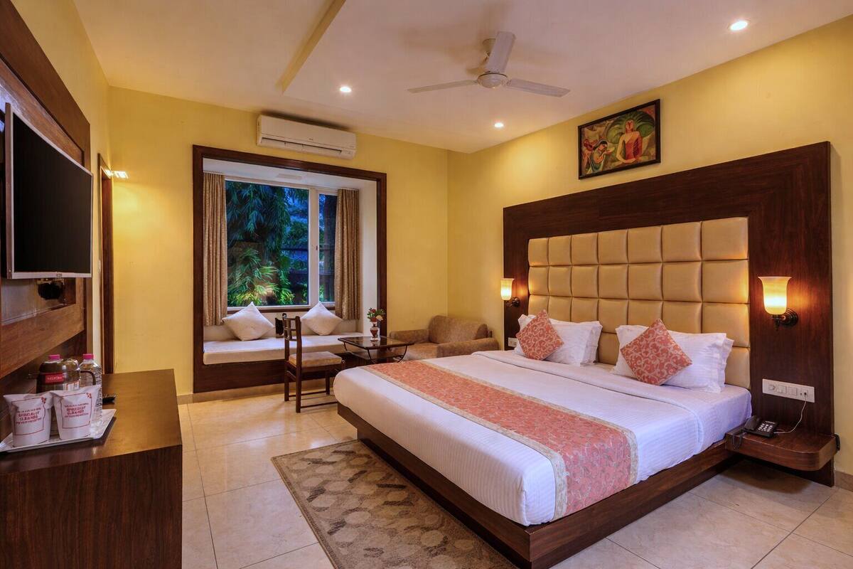 A hotel room with a large bed, a TV, a window with a view of greenery, a ceiling fan, and a painting on the wall.