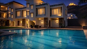 A multi-story hotel with a swimming pool, outdoor seating, and a view of a hilltop structure. - Devi Palace Monsoon Palace View Resort Udaipur (Udaipur)