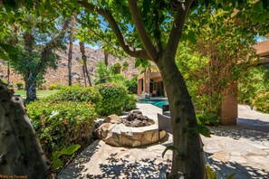 Surrounded by lush greenery and flowering plants, this serene garden features a stone fire pit and leads to a pool area.