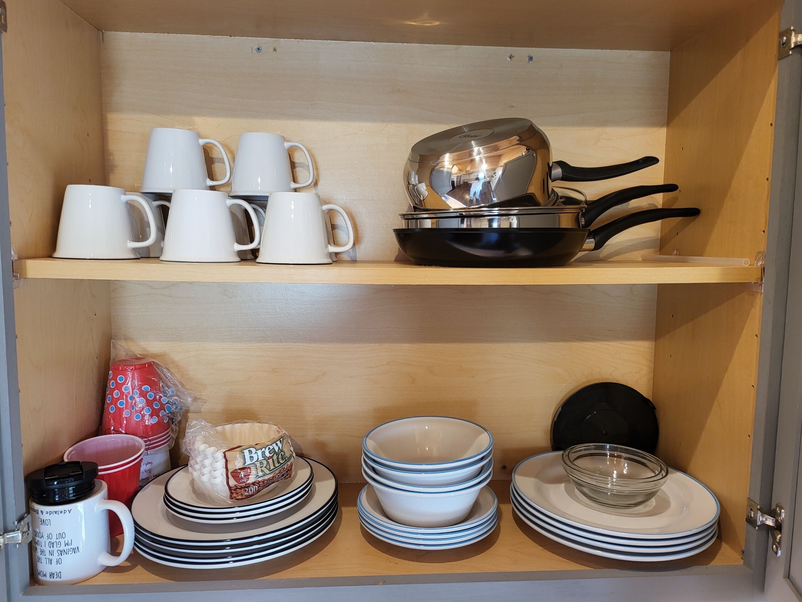 A kitchen cabinet with white mugs, a frying pan, and stacked plates and bowls.