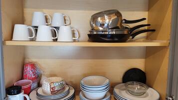 A kitchen cabinet with white mugs, a frying pan, and stacked plates and bowls.