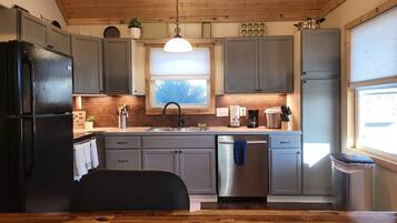 A kitchen with wooden cabinets, a black refrigerator, a stainless steel dishwasher, and a window with a view of trees.