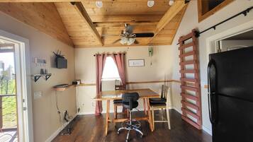 A compact kitchen with wooden beams, a dining table with chairs, a black refrigerator, and a window with curtains.