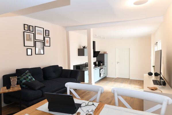 Ein modernes Wohnzimmer mit einer schwarzen Couch, einem Esstisch mit einem Laptop und einer Küche im Hintergrund.