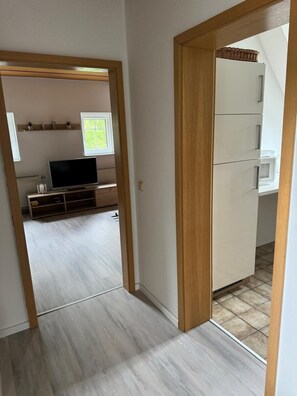 Ein Zimmer mit Fernseher, einer Ablage mit kleinen Pflanzen, einem Fenster und einer Tür zu einem weiteren Raum.