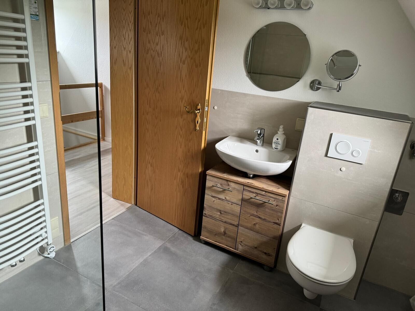 A bathroom with a wooden wardrobe, a white sink, a round mirror, and a toilet.