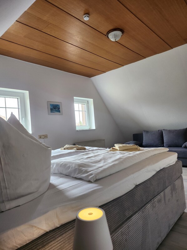 Ein Schlafzimmer mit schrägverdeckter Decke, zwei Fenstern, einem Bett, einem Nachttisch und einer Couch.