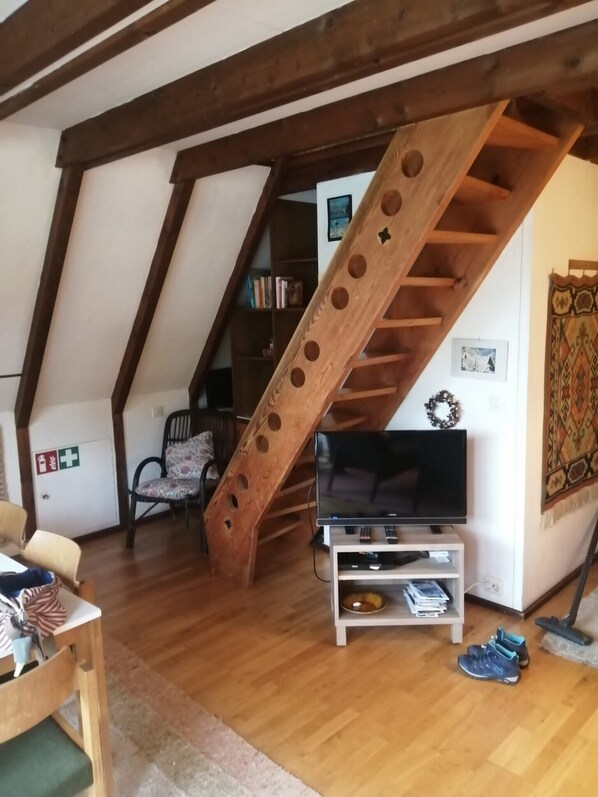 A loft-style living room with exposed wooden beams, a wooden staircase, a television on a white stand, a bookshelf, and a patterned wall hanging.