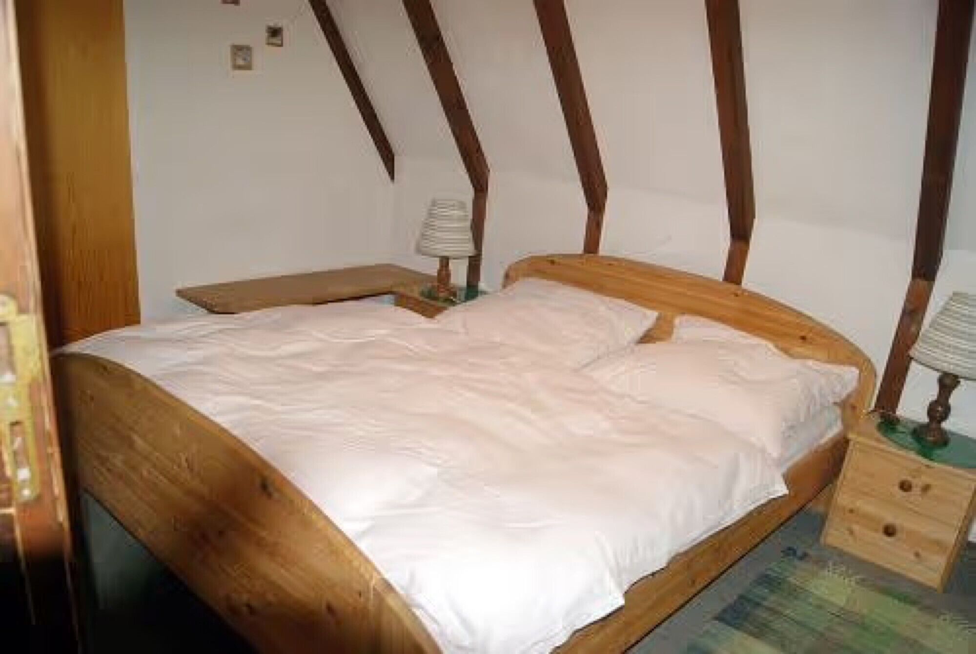 Ein Bett mit Holzbettgestell und weißer Bettwäsche in einem Raum mit schrägen Wänden und einem kleinen Nachttisch mit Lampe.
