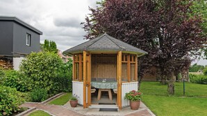 Ein hölzernes Gartenhaus mit Tisch und Stühlen, umgeben von einem Backsteinweg und Grünpflanzen.