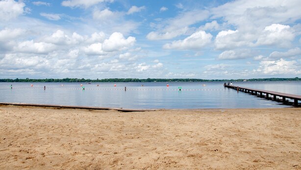 Ein Strand mit einem hölzernen Steg, der sich über ruhiges Wasser erstreckt, bewölkter Himmel und eine entfernte Küste.