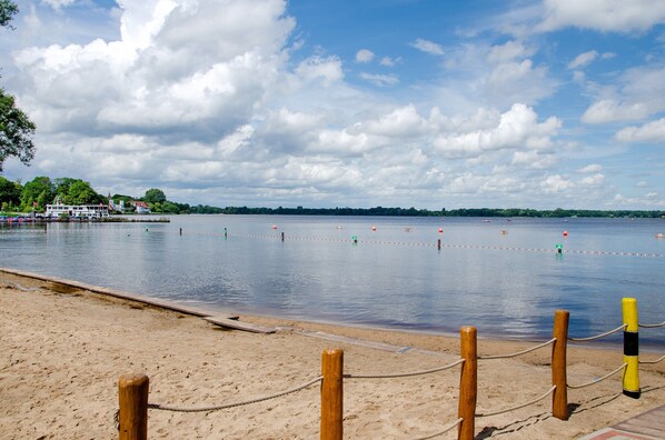 A beach with a wooden fence, a dock with boats, and a calm lake under a partly cloudy sky.