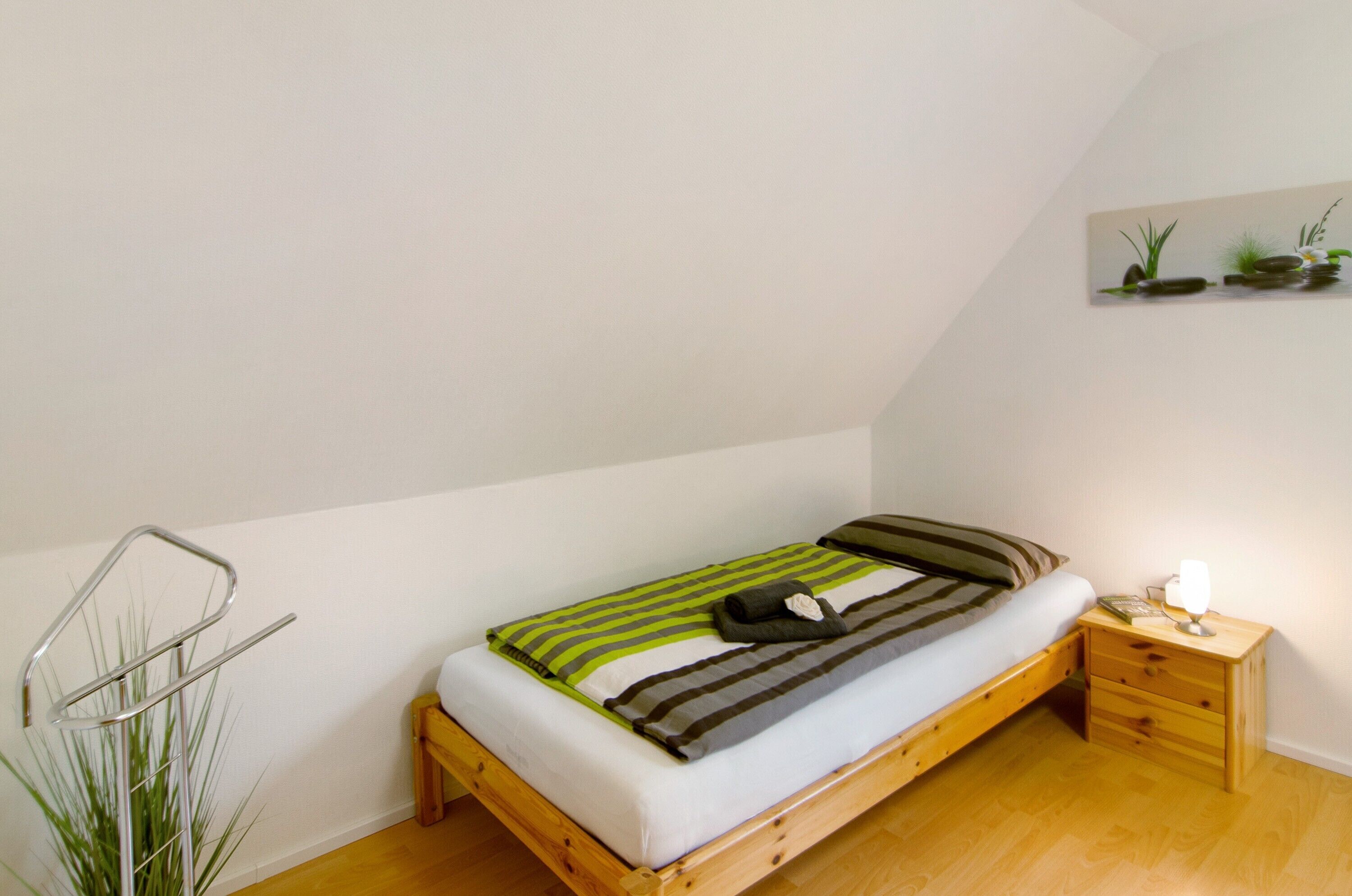 A single bed with a wooden frame, a bedside table with a lamp, and a wall-mounted shelf with plants.