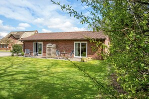 Un bâtiment en briques surmonté d’un toit en tuiles, avec une pelouse et une terrasse aménagée avec du mobilier d’extérieur.