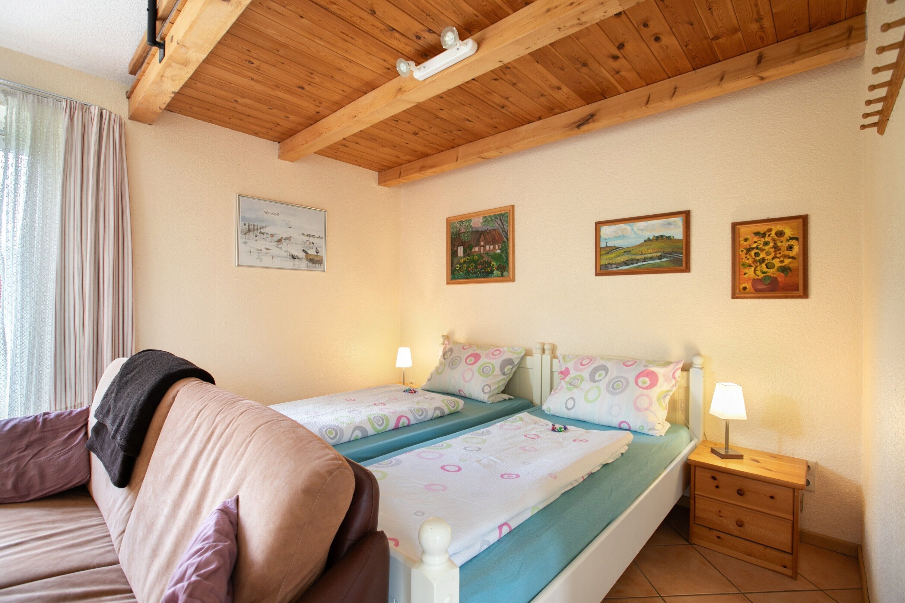 A bedroom with a wooden ceiling, two beds with patterned bedding, a nightstand with a lamp, and wall art.
