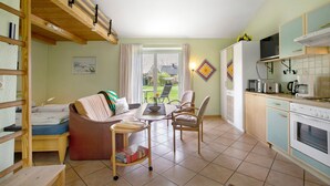 A compact living space with a sofa, chairs, a small table, a kitchen area with a stove and microwave, and a window with curtains. - Apartment 'Eulenhof 1' with Private Terrace, Private Garden and Wi-Fi (Bordelum)