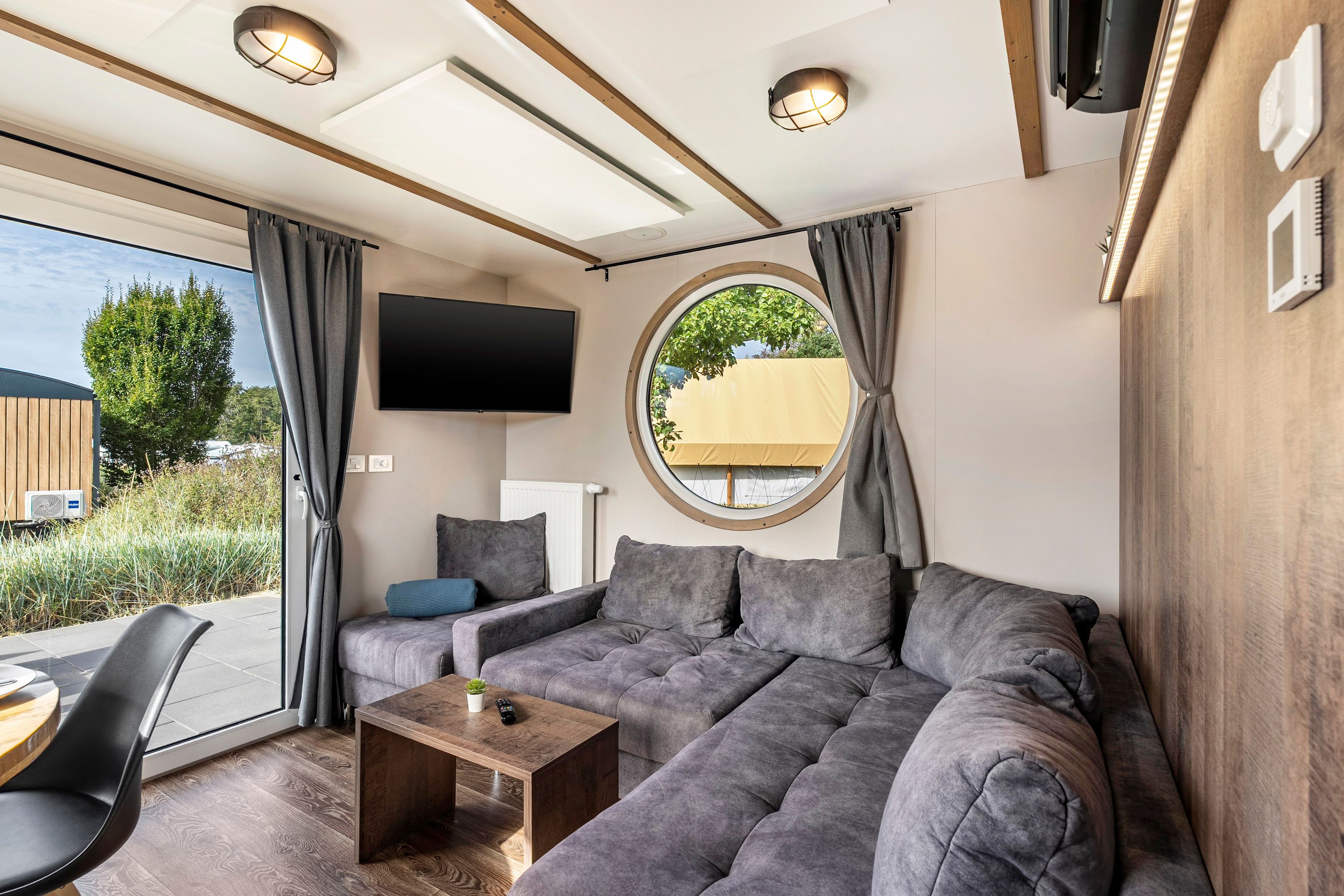 Ein modernes Wohnzimmer mit einem Ecksofa, einem Couchtisch, einem an der Wand montierten Fernseher und einem großen Fenster mit Vorhängen.