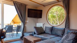 A modern living area with a sofa, a coffee table, and a television. There is a window with a view of trees and a tent outside. - Holiday Home 'Kaperfahrt' with Private Terrace, Wi-Fi and Air Conditioning (Großenbrode)