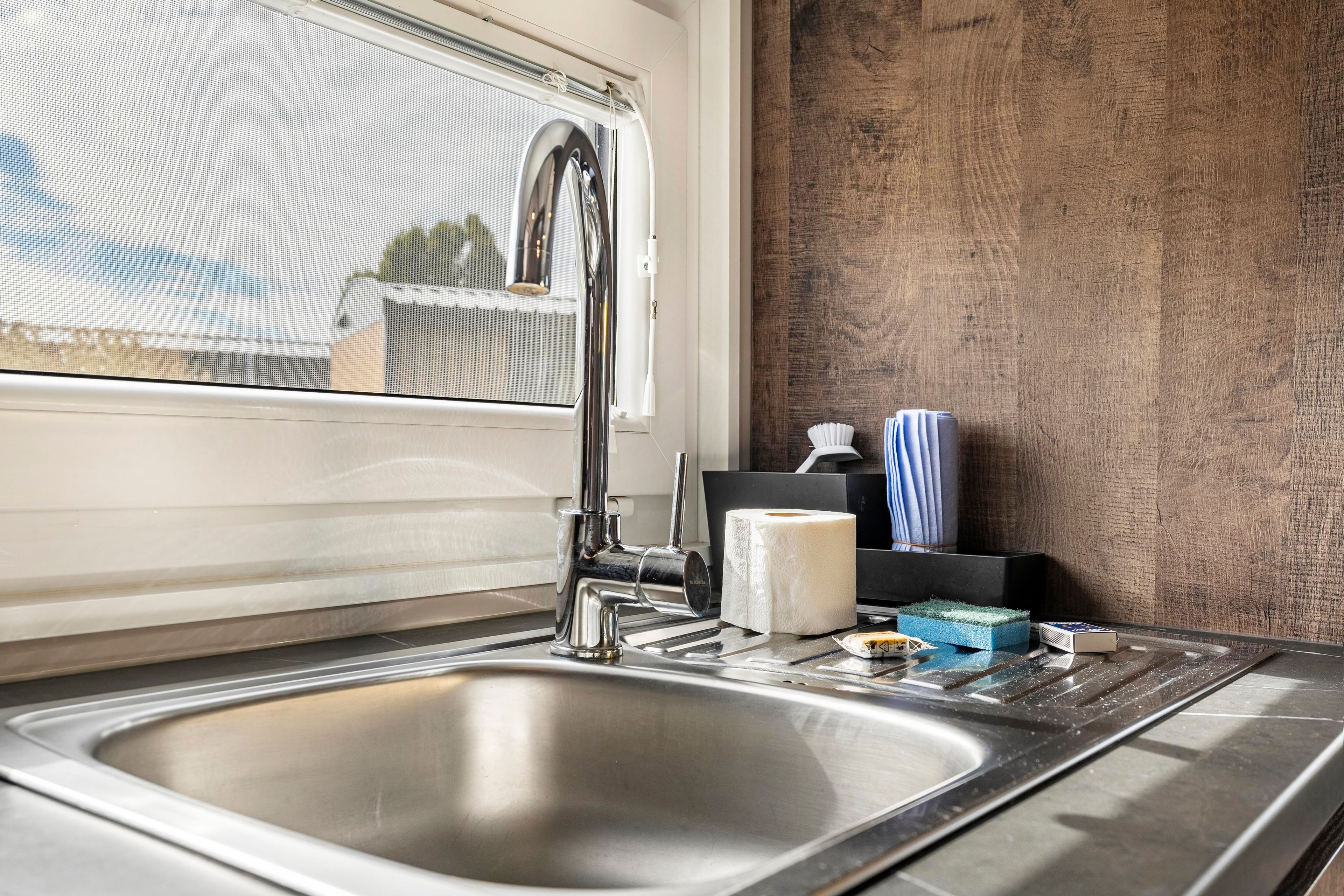 Eine moderne Küche mit Spüle aus Edelstahl, einem Wasserhahn und einem Fenster mit Blick ins Freie.
