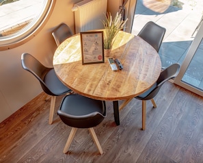 A round wooden table with a menu, surrounded by four chairs, in a room with a large window and a radiator. - Holiday Home 'Kaperfahrt' with Private Terrace, Wi-Fi and Air Conditioning (Großenbrode)