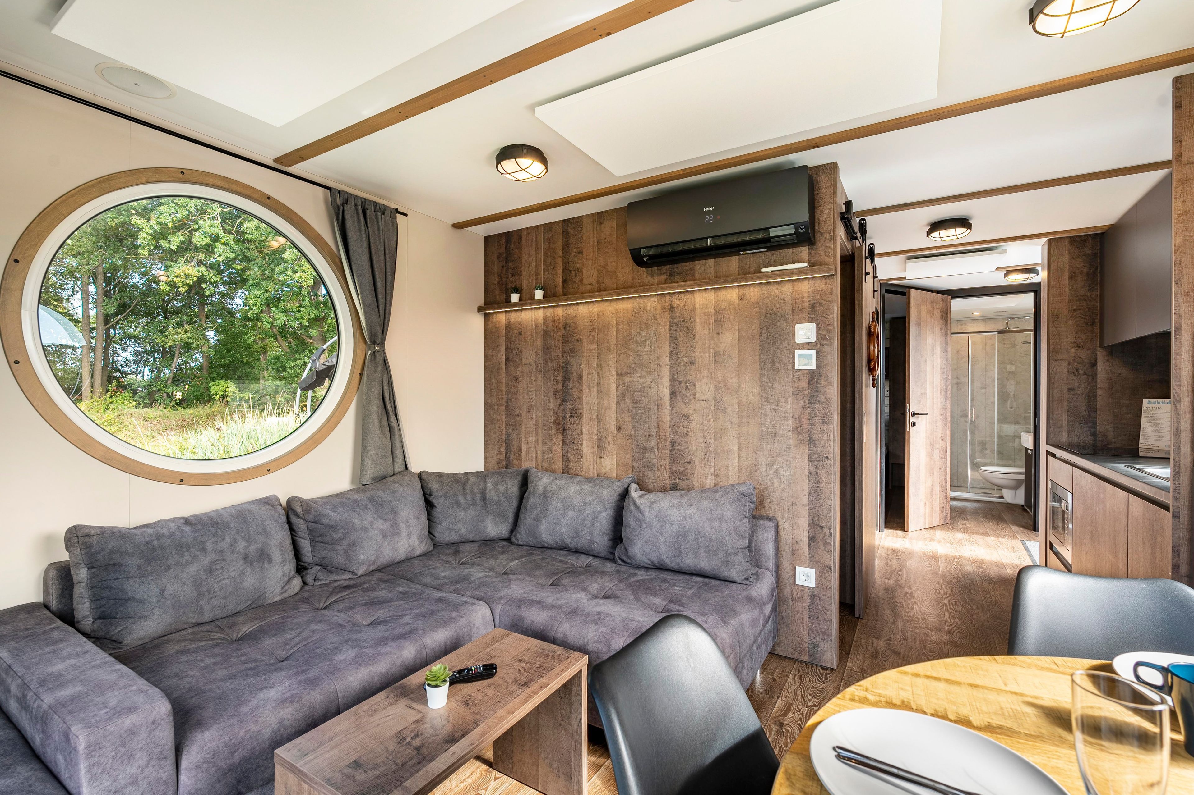 Een modern interieur van een camper met een modulaire bank, een eettafeltje en een rond raam met uitzicht op bomen.