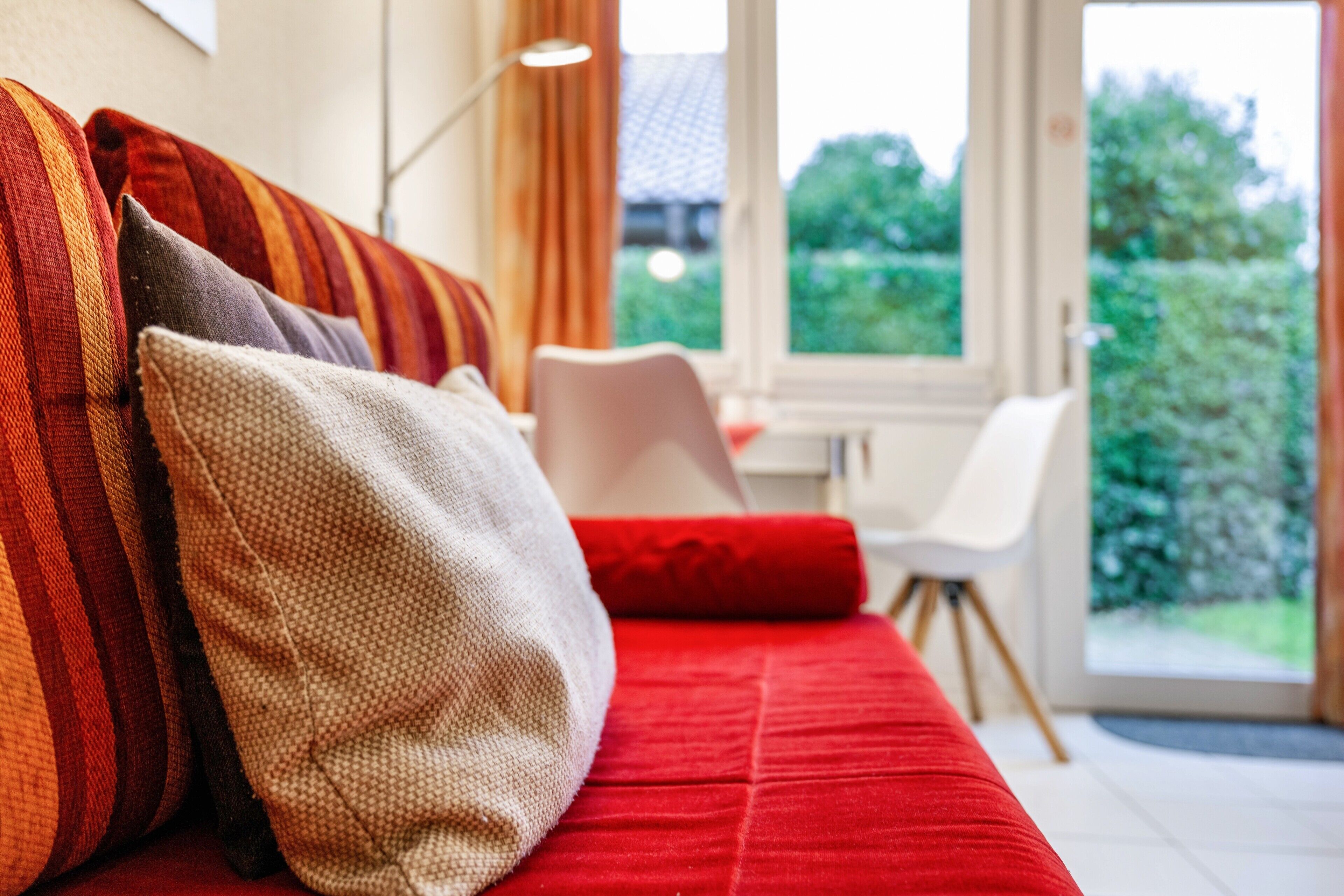A red sofa with patterned cushions, a white chair, and a view of greenery outside.