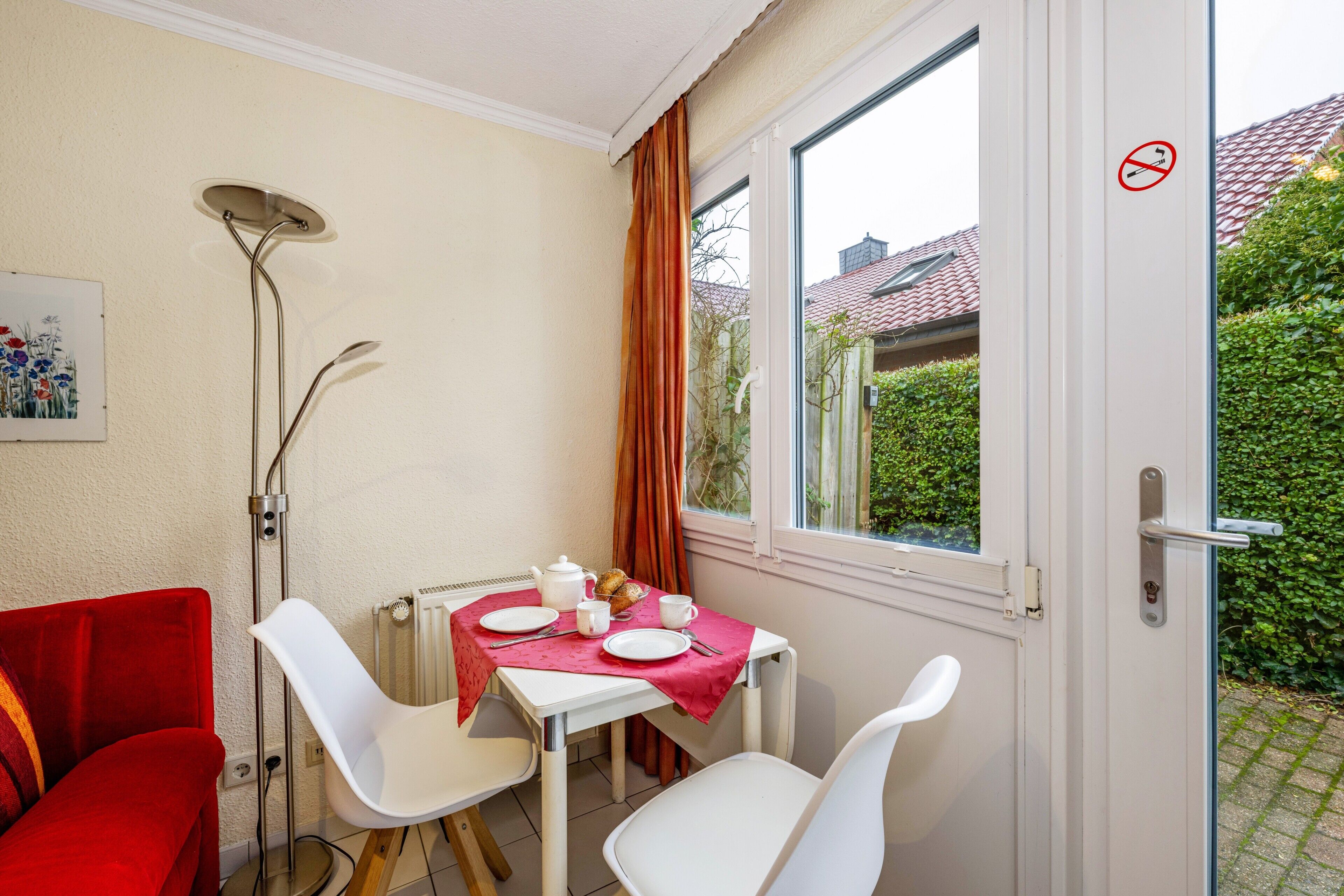 A room with a dining table set for two, a radiator, a window with curtains, and a red sofa.