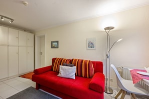 Un canapé rouge avec des coussins, un radiateur blanc, une chaise blanche, un lampadaire et une porte donnant sur une armoire blanche.