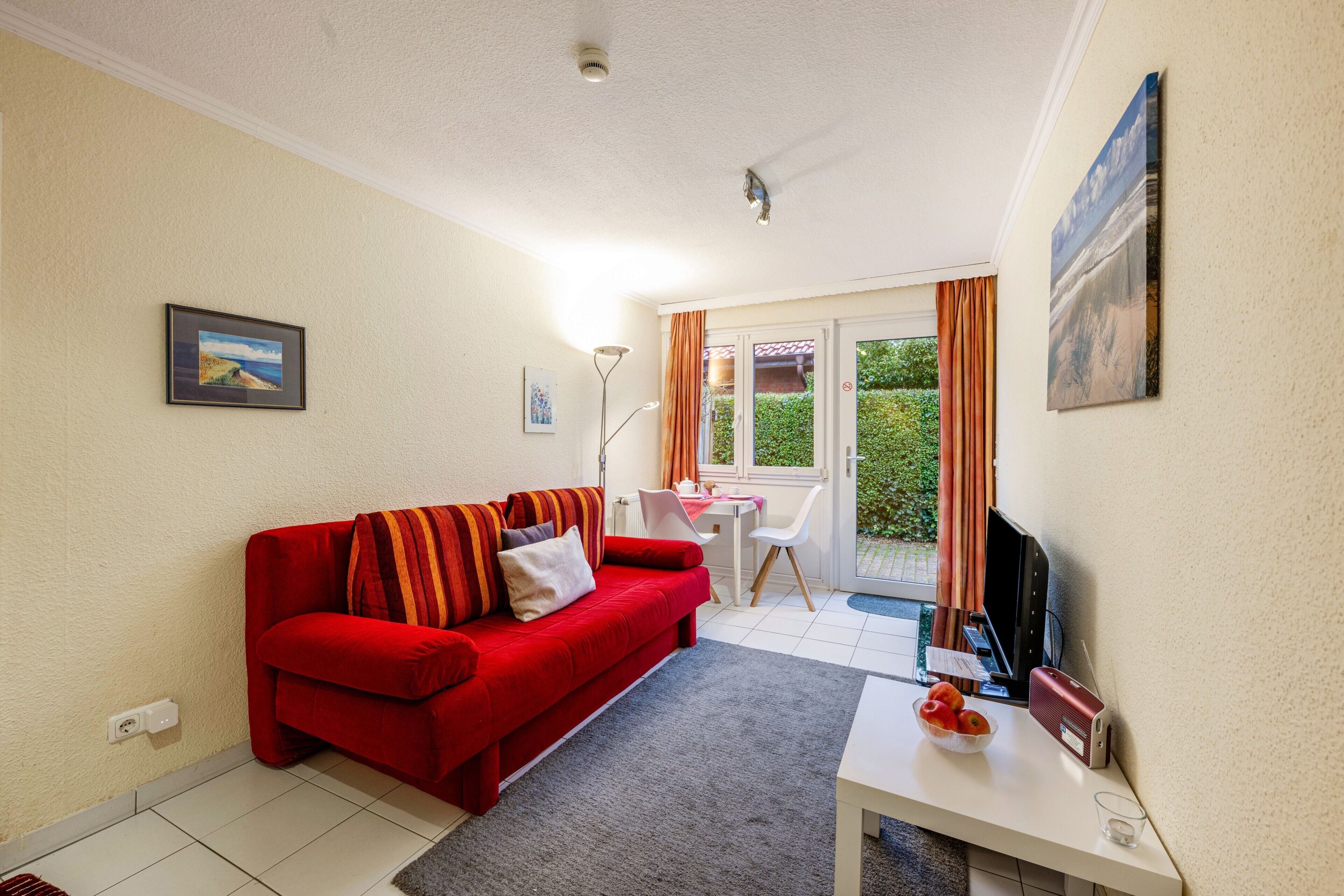 A living room with a red sofa, a white table, a flat-screen TV, and a window with curtains.