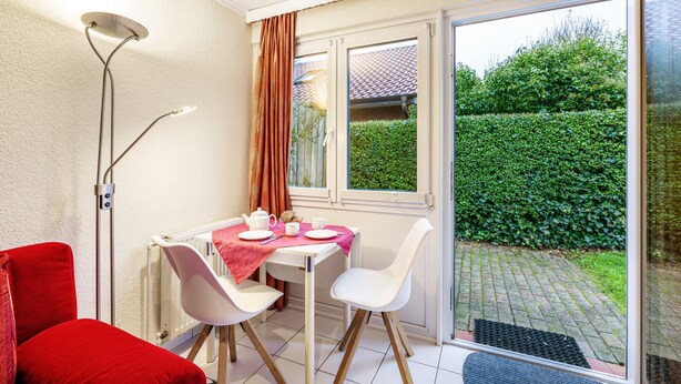 Une pièce comprenant une table, des chaises, un radiateur, une fenêtre avec des rideaux et une vue sur la verdure à l’extérieur.