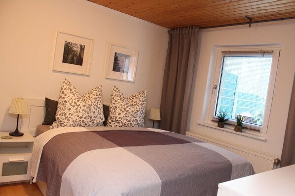 Une chambre à coucher comprenant un lit, deux tables de chevet, une lampe, une fenêtre avec des stores et deux tableaux encadrés accrochés au mur.