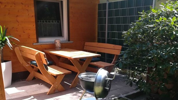 Een terras buiten met een houten tafel en stoelen, een barbecue en een bloempot.