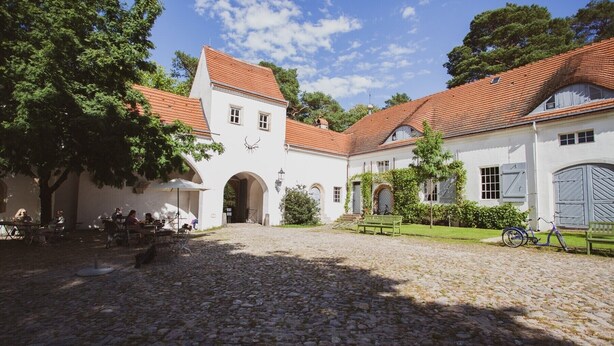 Ein Innenhof mit Kopfsteinpflaster, weißen Gebäuden mit roten Ziegeldächern und einer überdachten Sitzgelegenheit mit Tischen und Stühlen.