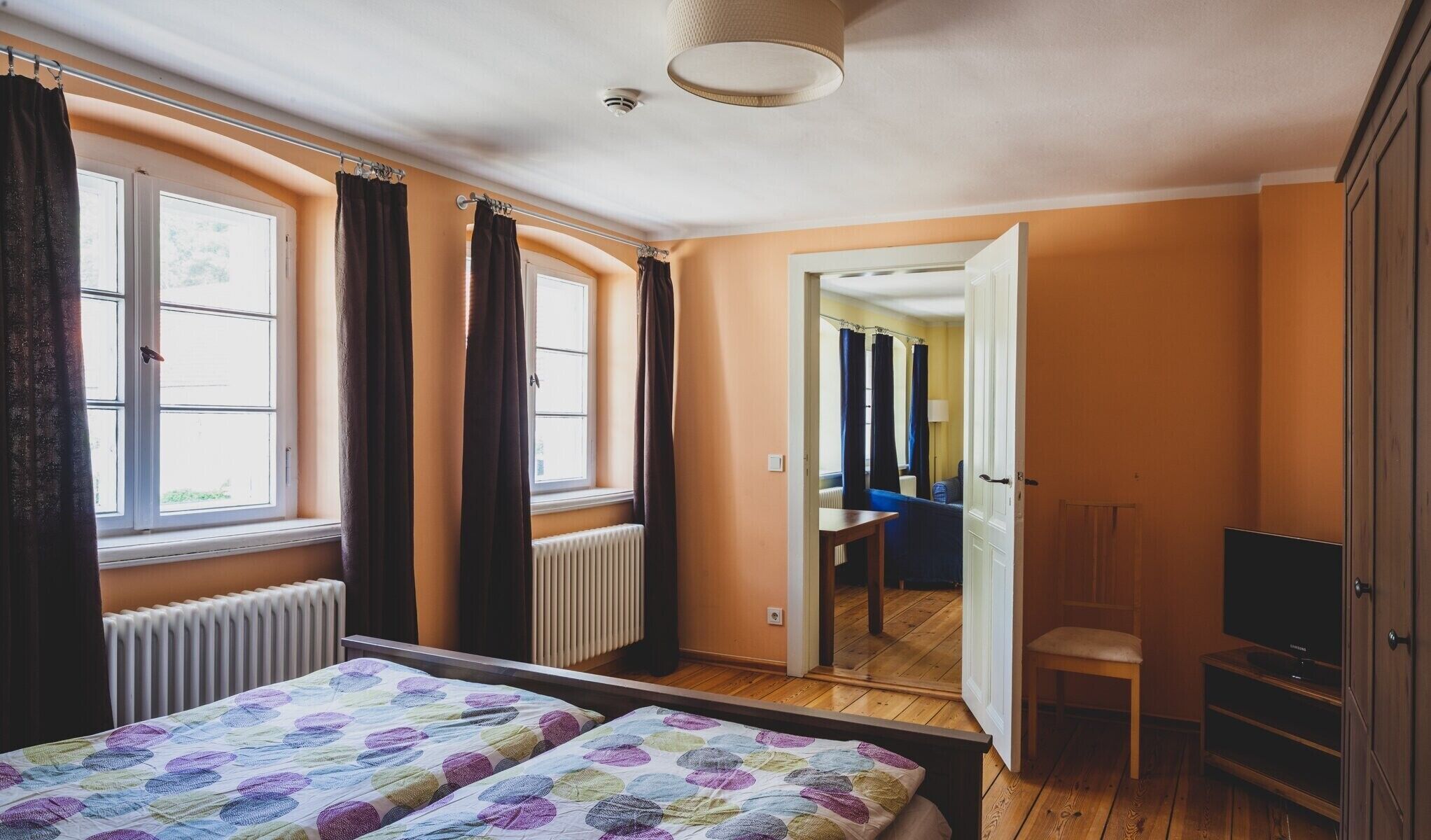 A bedroom with a bed, a chair, a TV, and a view of another room.
