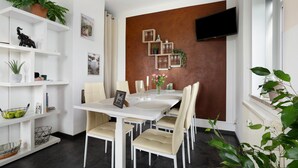 A dining area with a white table, beige chairs, a TV mounted on a brown wall, and shelves with plants and decorative items.