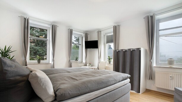 A bedroom with a bed, a TV, a window with curtains, and a radiator.