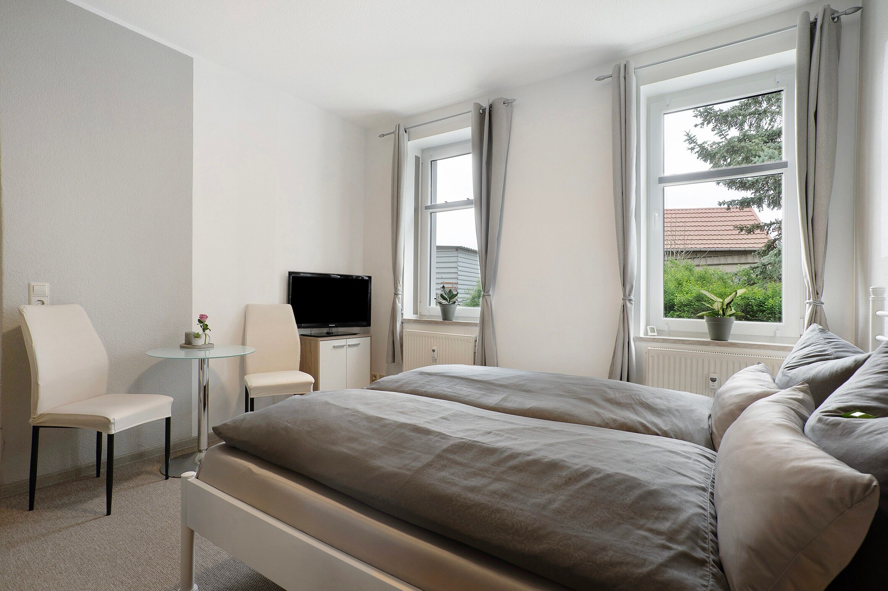 Ein Hotelzimmer mit einem Bett, zwei Stühlen, einem kleinen Tisch mit Fernseher und einem Fenster mit Vorhängen.