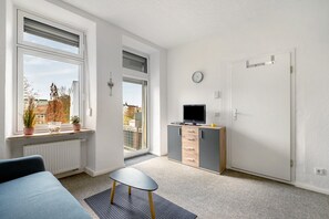 A room with a bed, a TV on a cabinet, a window with blinds, a door, and a view of buildings outside.