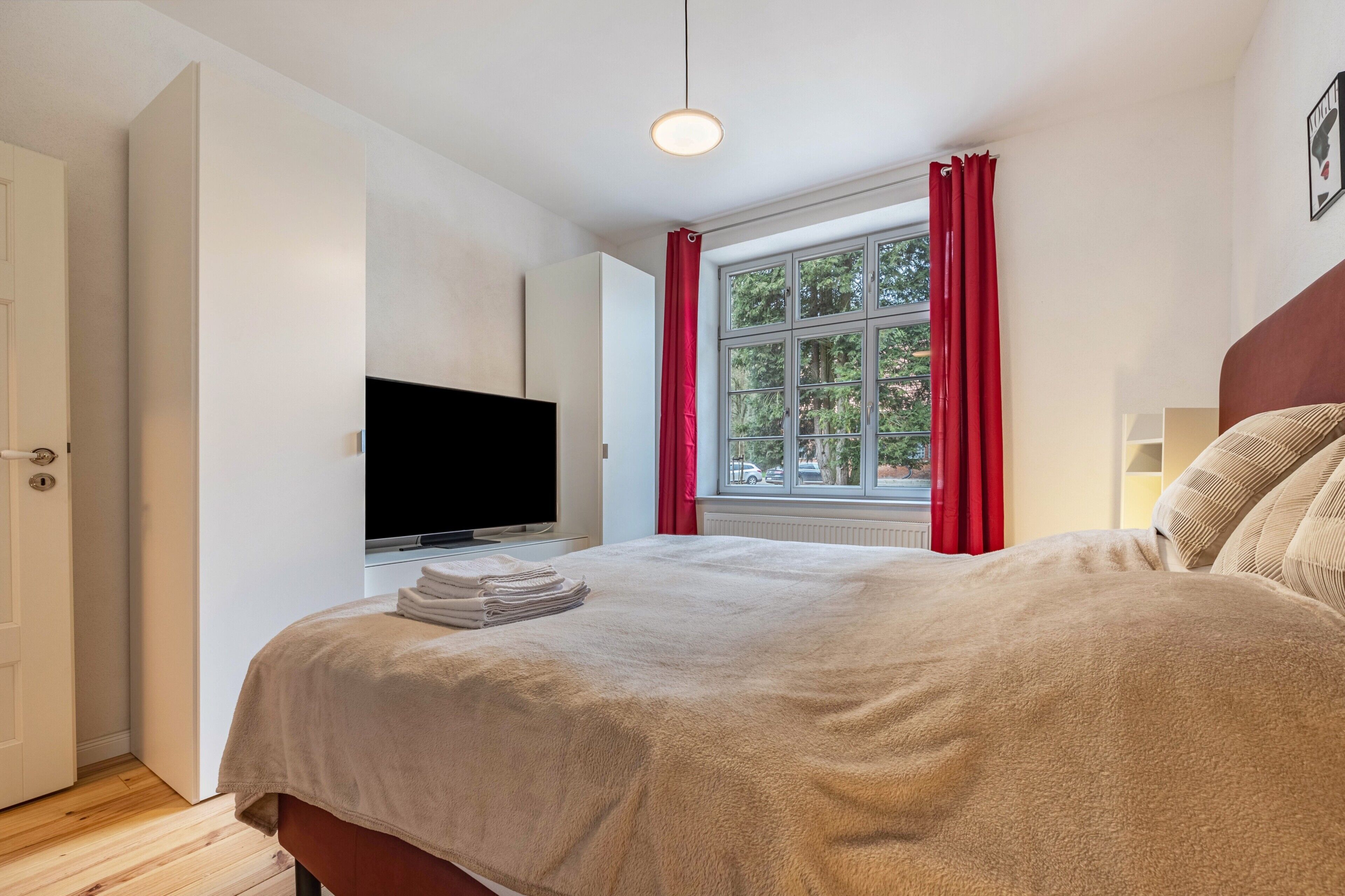 Ein Schlafzimmer mit einem Bett, einem Fernseher, einem Fenster mit roten Vorhängen und einer Tür.