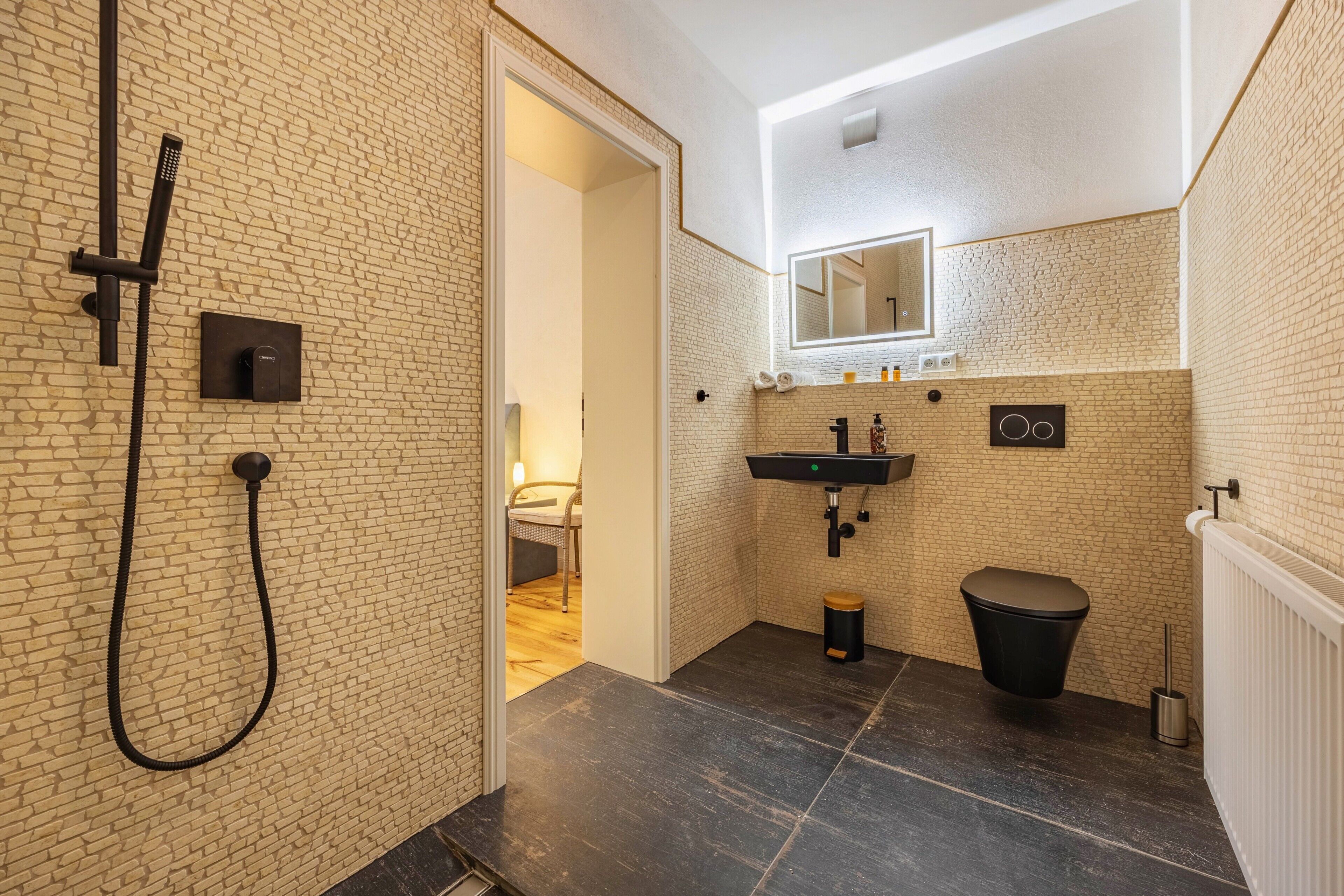 Een moderne badkamer met douche, wastafel en toilet, met een metselwerk wand en een zichtbare deur naar een andere kamer.