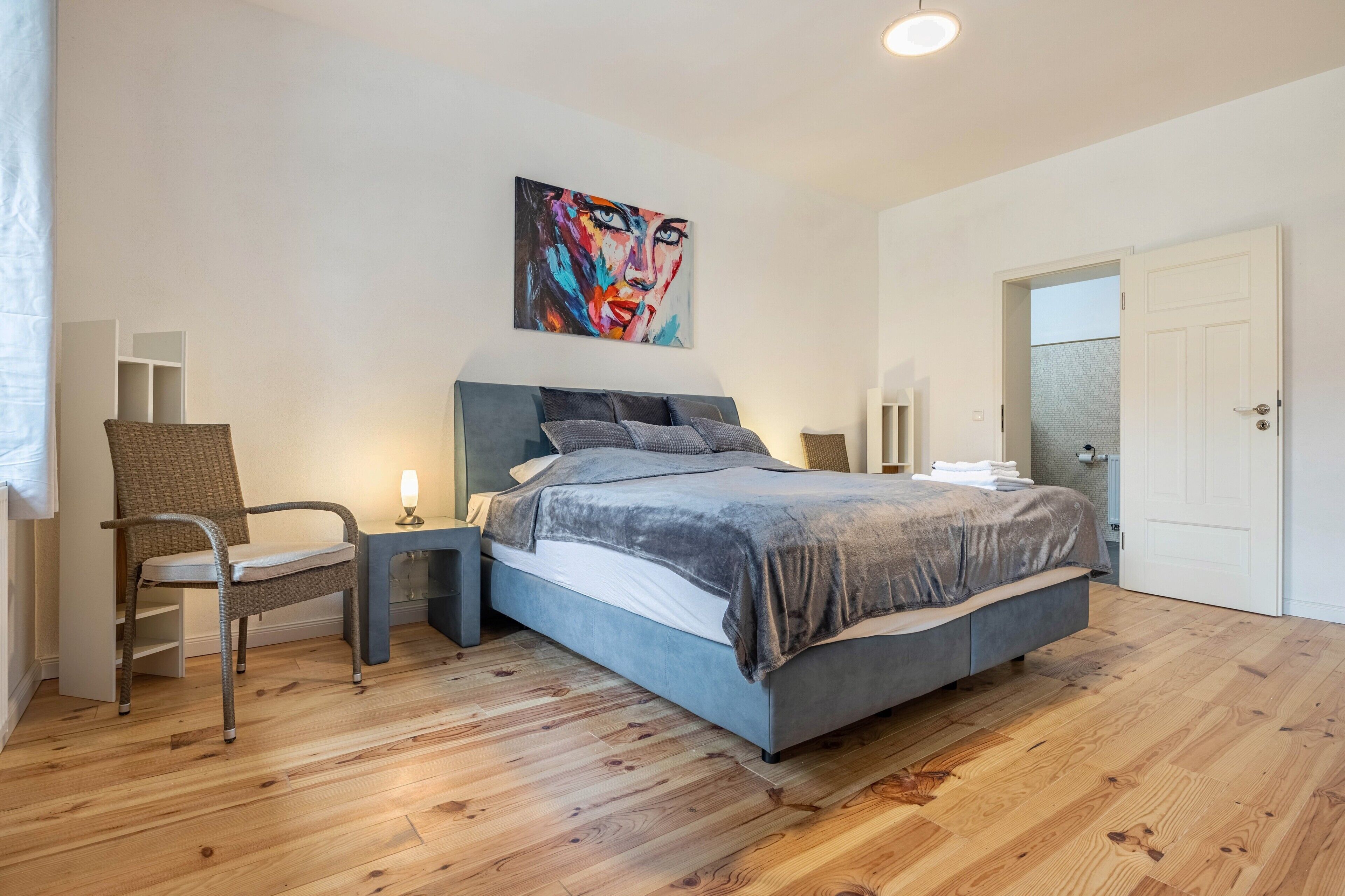 A bedroom with a bed, a chair, a bedside table, a wall-mounted artwork, and a door.