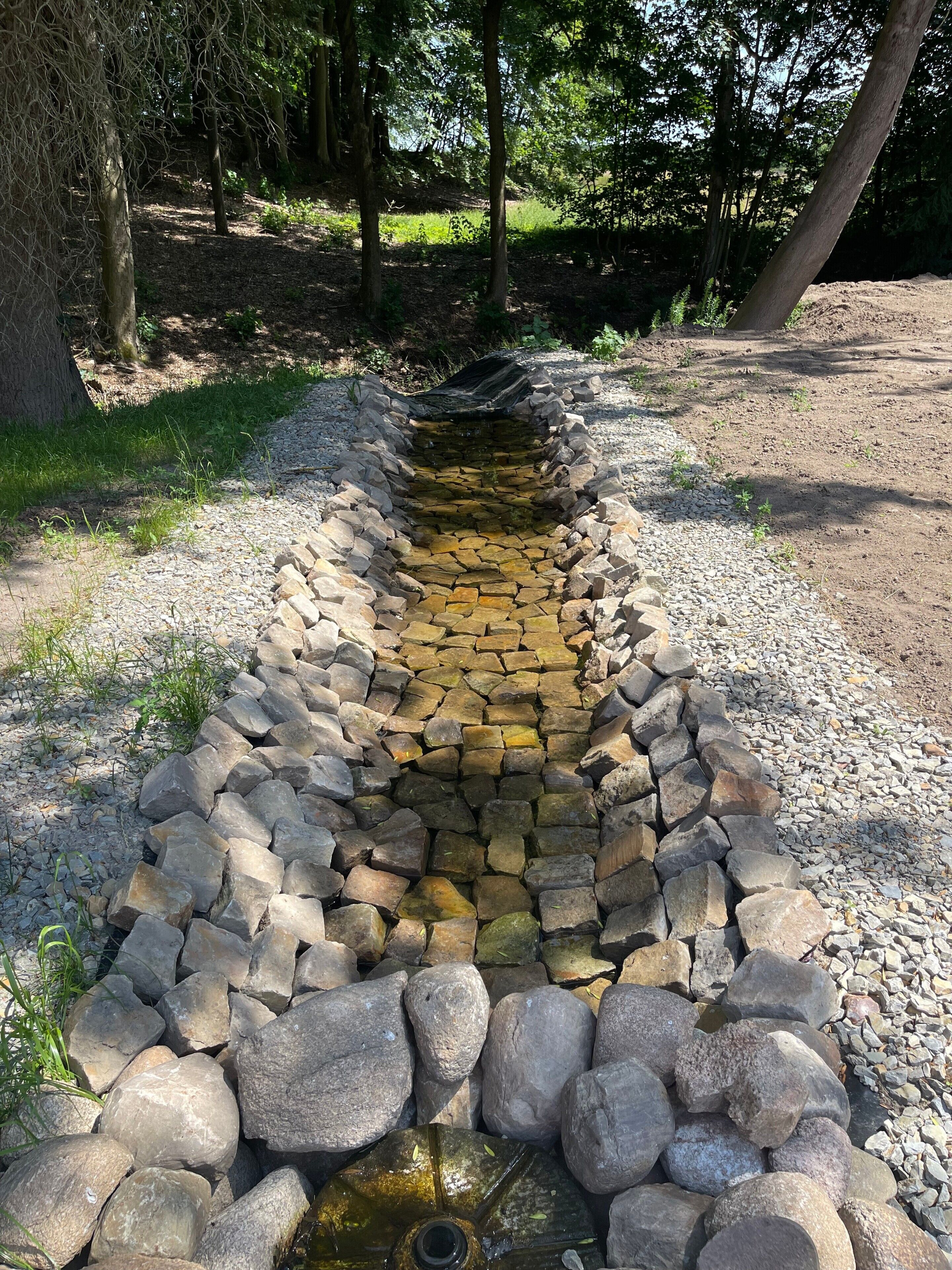 En stenvandinstallation med en smal strøm, omgivet af sten og grus, i en skovpark.