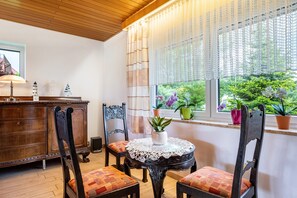 A room with a wooden ceiling, a window with a view of greenery, a wooden cabinet, and a round table with a lace tablecloth.