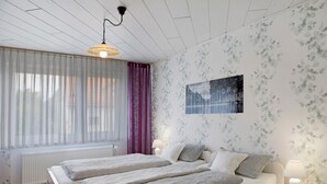 Et soveværelse med to senge, et vindue med lette gardiner, en radiator, et billed på væggen og en sengebordlampe.