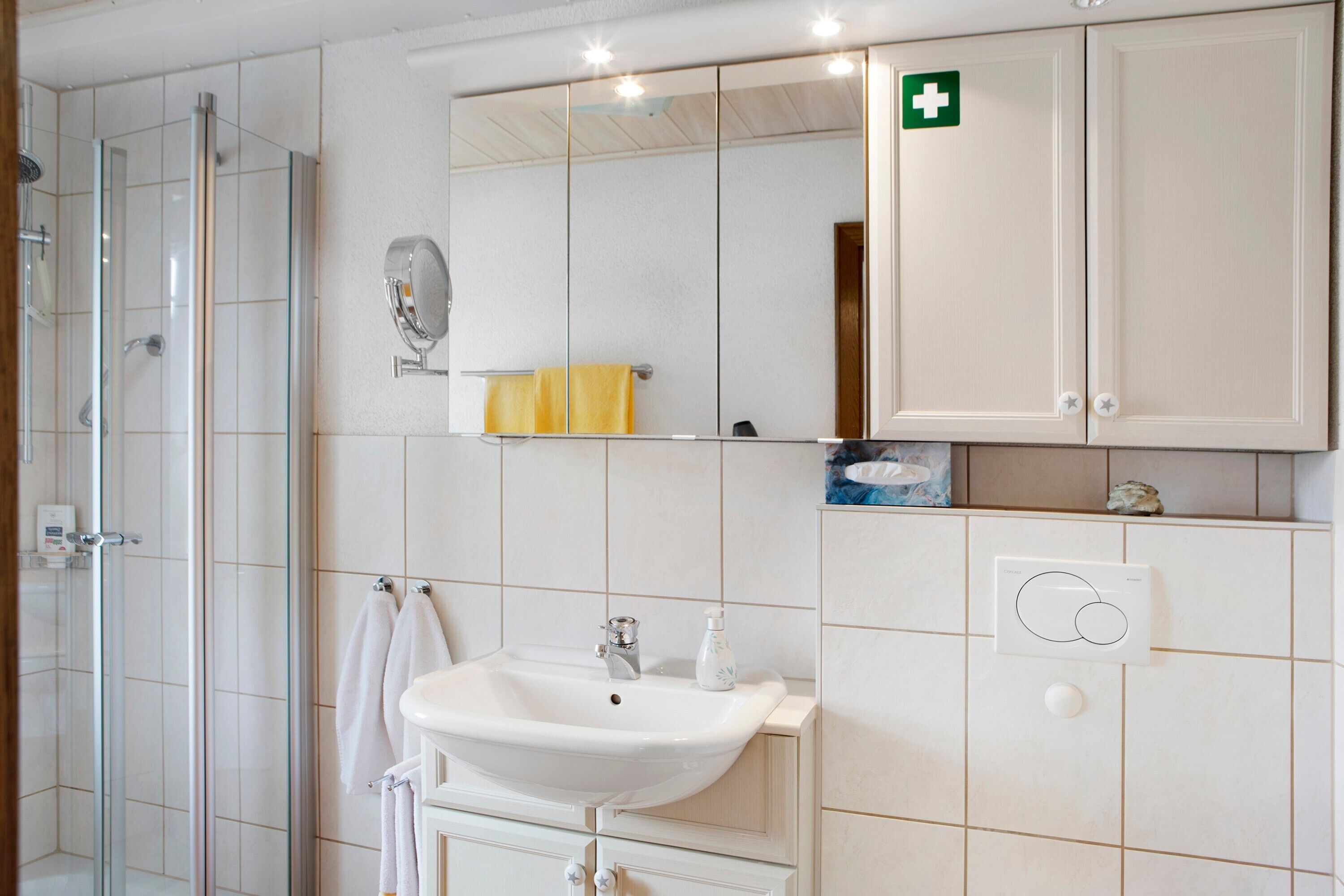 Une salle de bain avec une douche à parois de verre, un lavabo, un miroir et un meuble sur lequel figure le symbole des premiers secours.