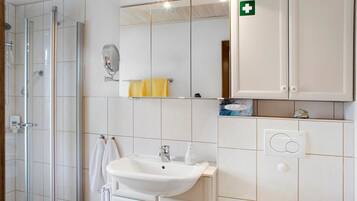 Une salle de bain avec une douche à parois de verre, un lavabo, un miroir et un meuble sur lequel figure le symbole des premiers secours.