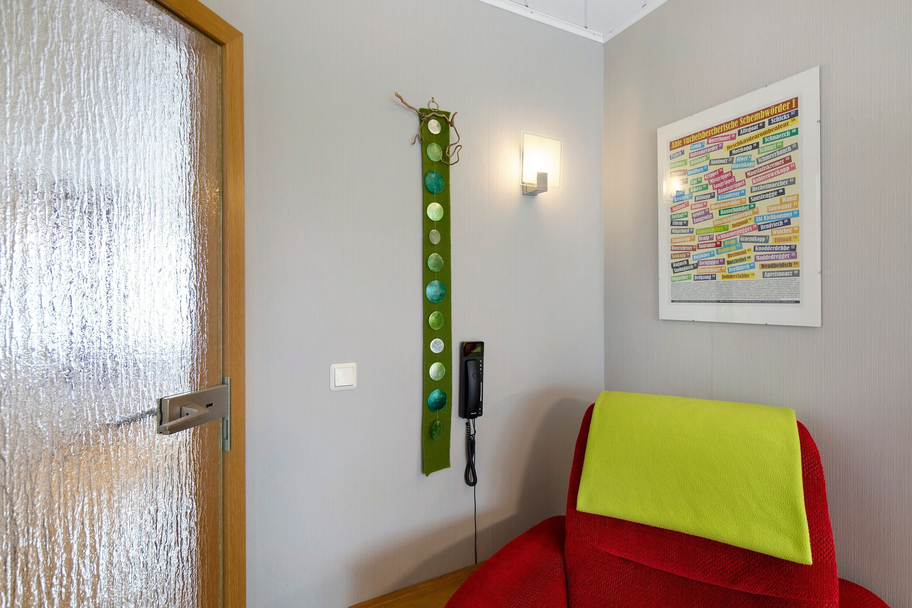 A room with a wooden door, a green wall decoration, a red chair, and a wall-mounted light.