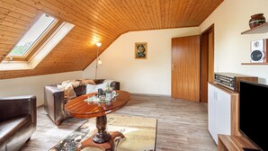 A loft-style room with a sloped ceiling, a round wooden dining table, a sofa, a television, and a small kitchen area.