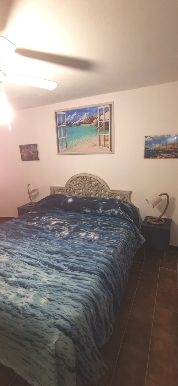 A bedroom with a bed, a ceiling fan, a nightstand with a lamp, and a wall with a window and two paintings.