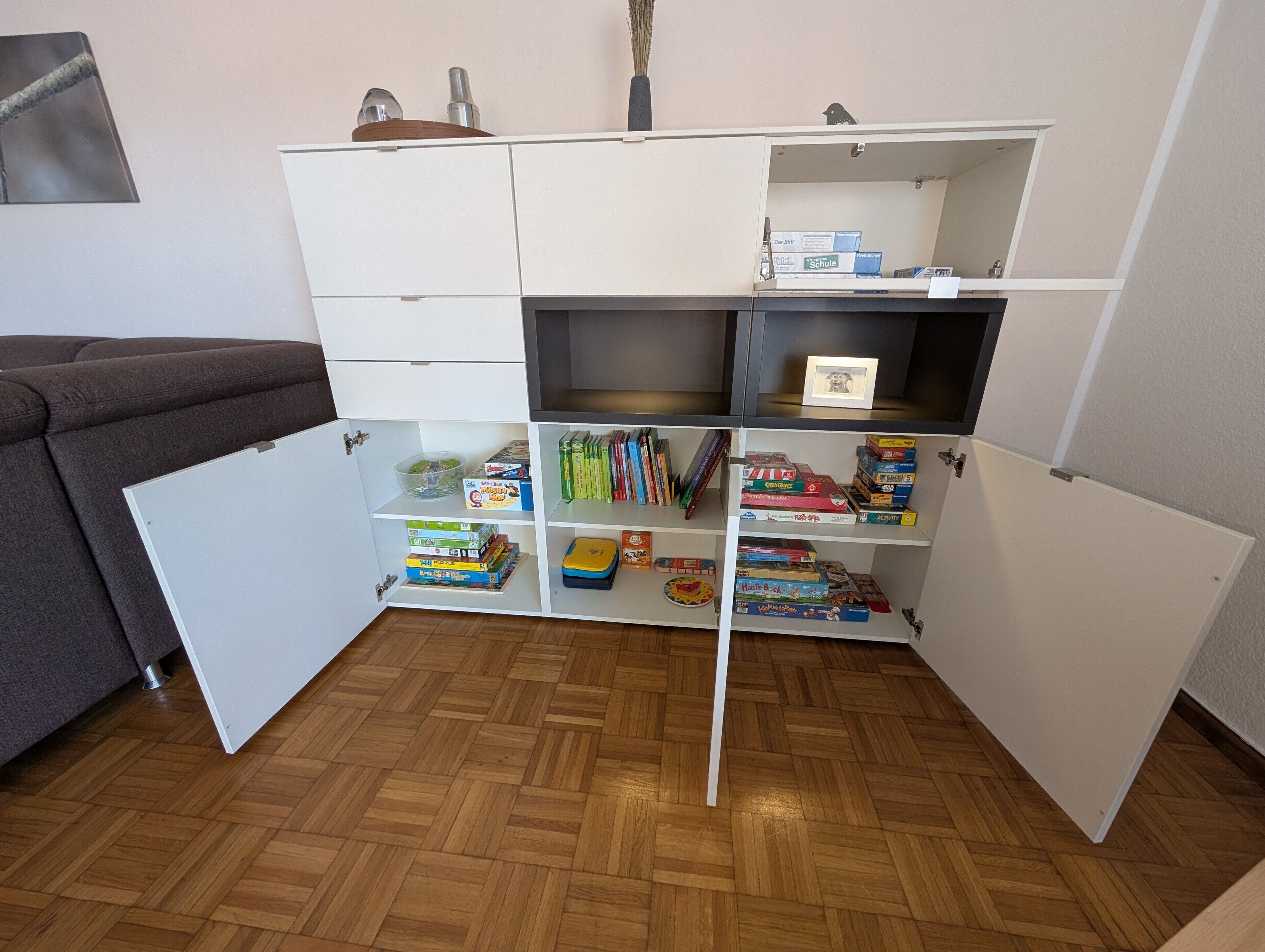 A room with a wooden herringbone floor, a white cabinet with open shelves containing books and board games, and a grey sofa.